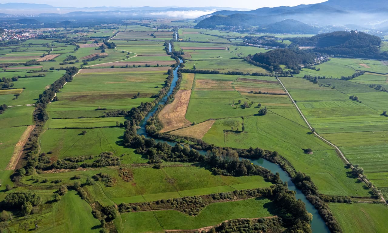 Vue aérienne d’une plaine agricole traversée par une rivière bordée d’arbres, collines à l’horizon